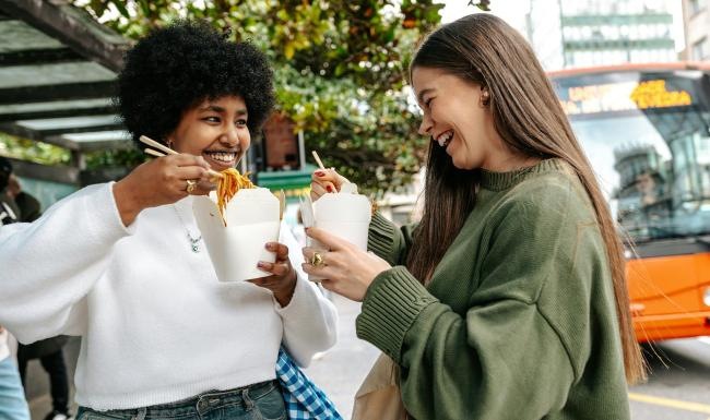Two women laughing and eating street food in the city.